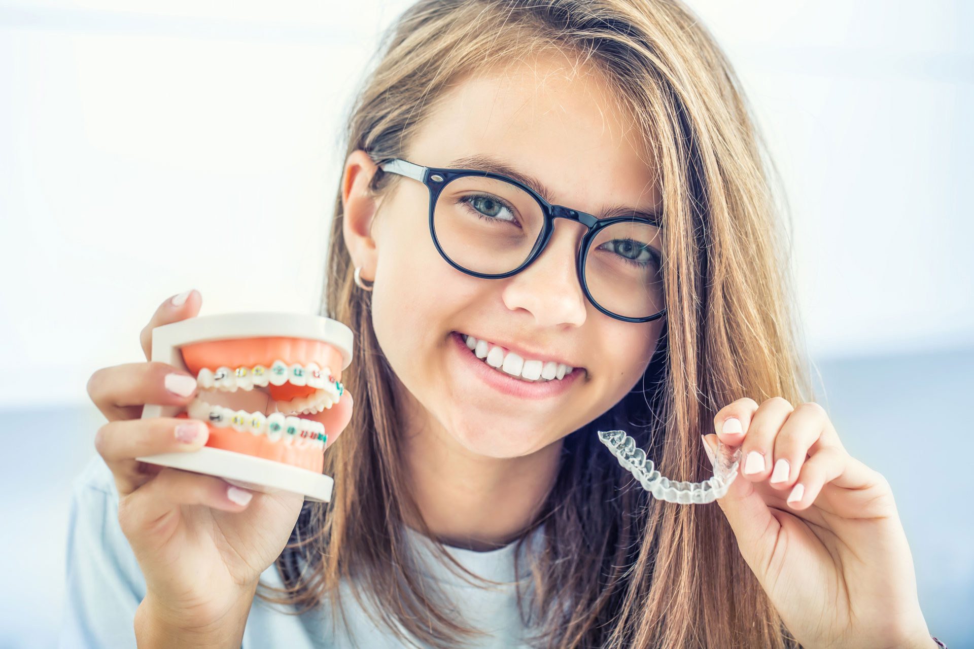 teen girl holding invisalign aligners in one hand and a tooth model showing braces in the other